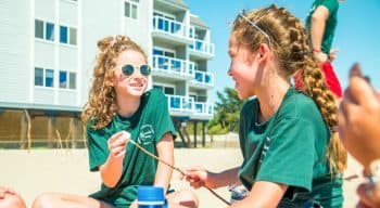 A girl with sunglasses talks to another girl with marshmallows on a stick