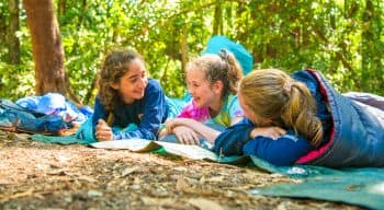 Three girls in sleeping bags laughing and relaxing on the forest floor