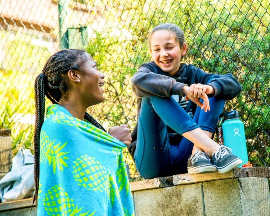 Girl with a towel talks to girl sitting on fence