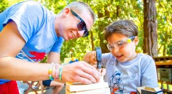 A little boy hammering wood with the help of a counselor