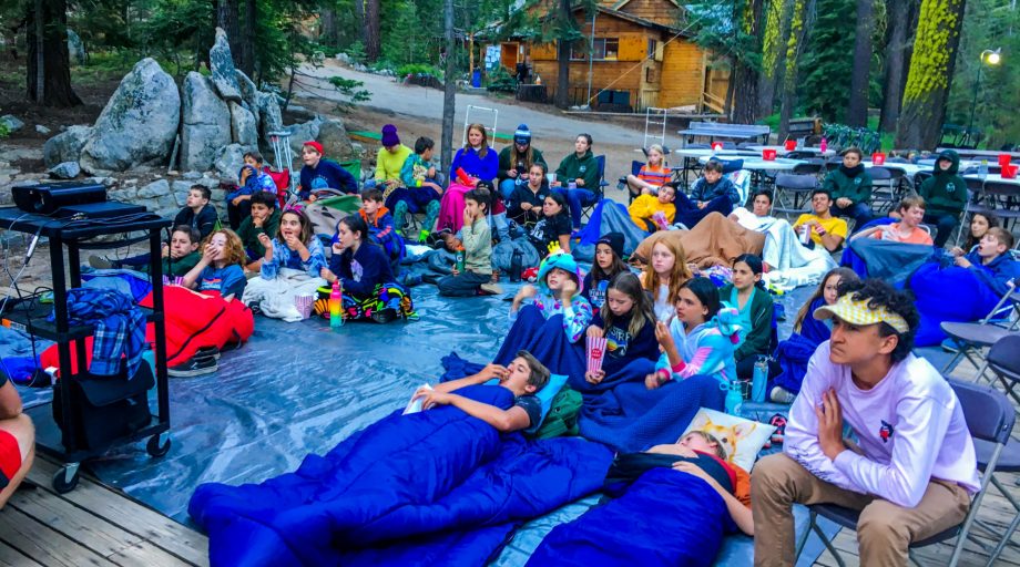 campers in sleeping bags sit outside to watch a movie